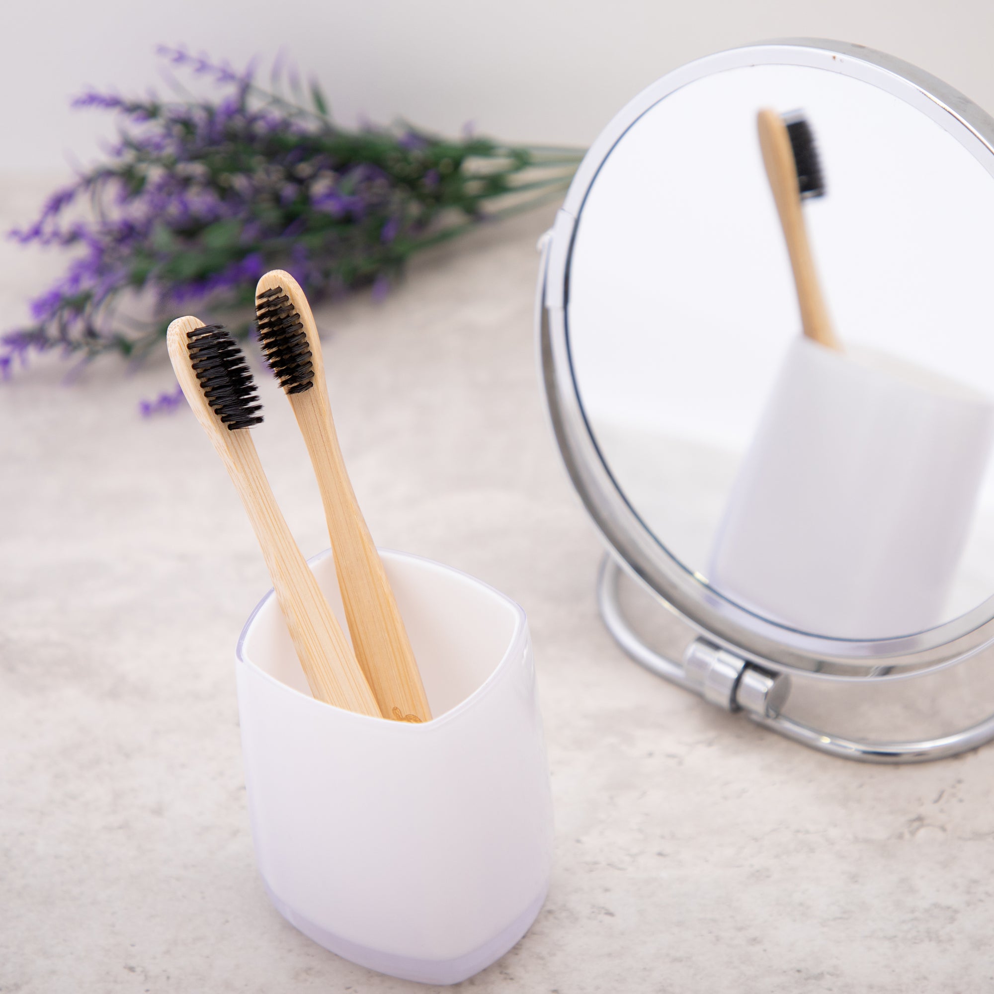 Two bamboo toothbrushes in a white holder with a mirror and lavender flowers in the background.
